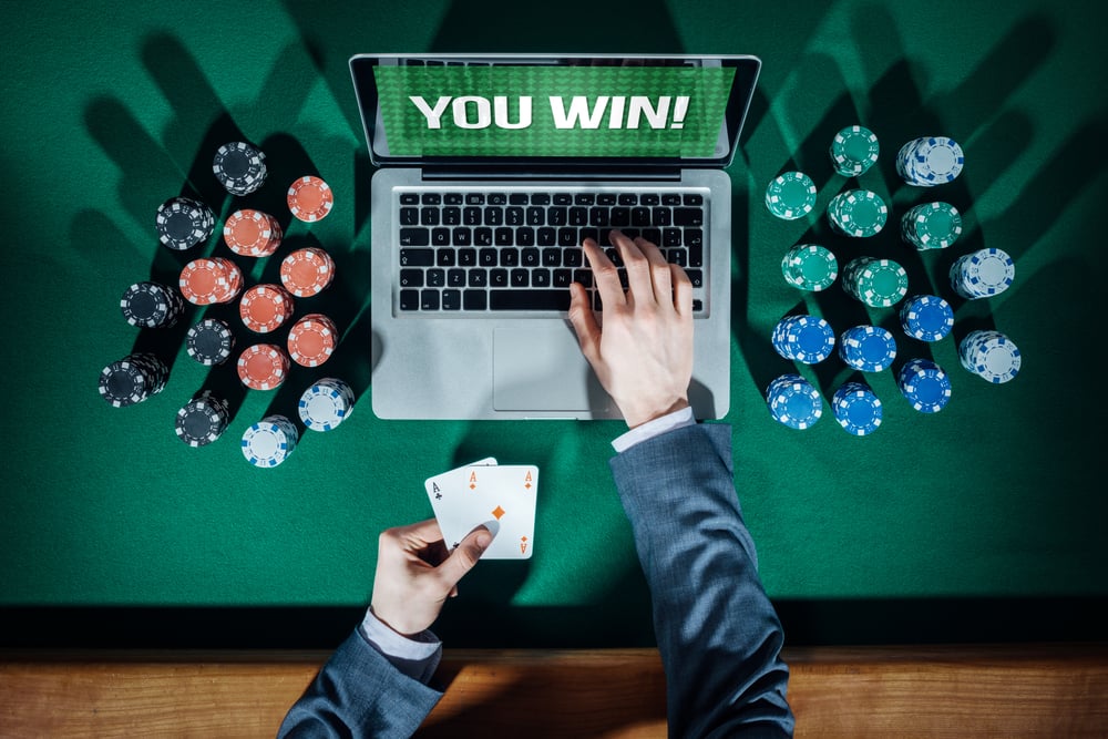 man in suit holds cards as he plays online poker on laptop surrounded by chips