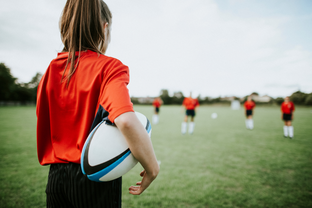 female rugby player holding football as she looks on at her team